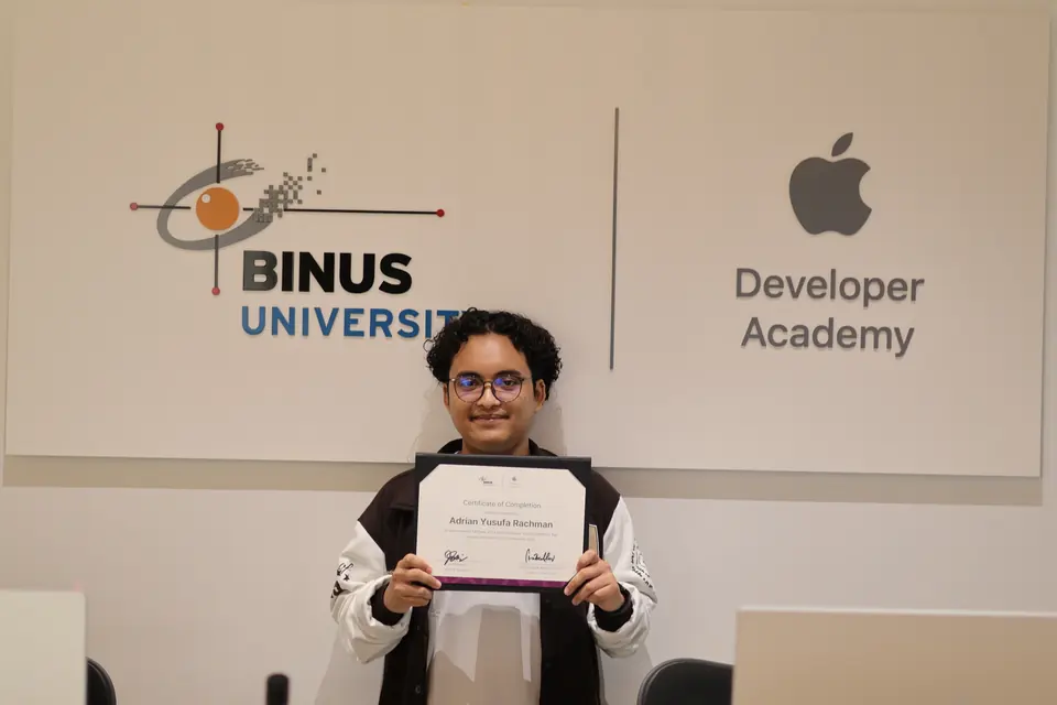 Adrian holding his graduation certificate in front of the BINUS and Developer Academy sign