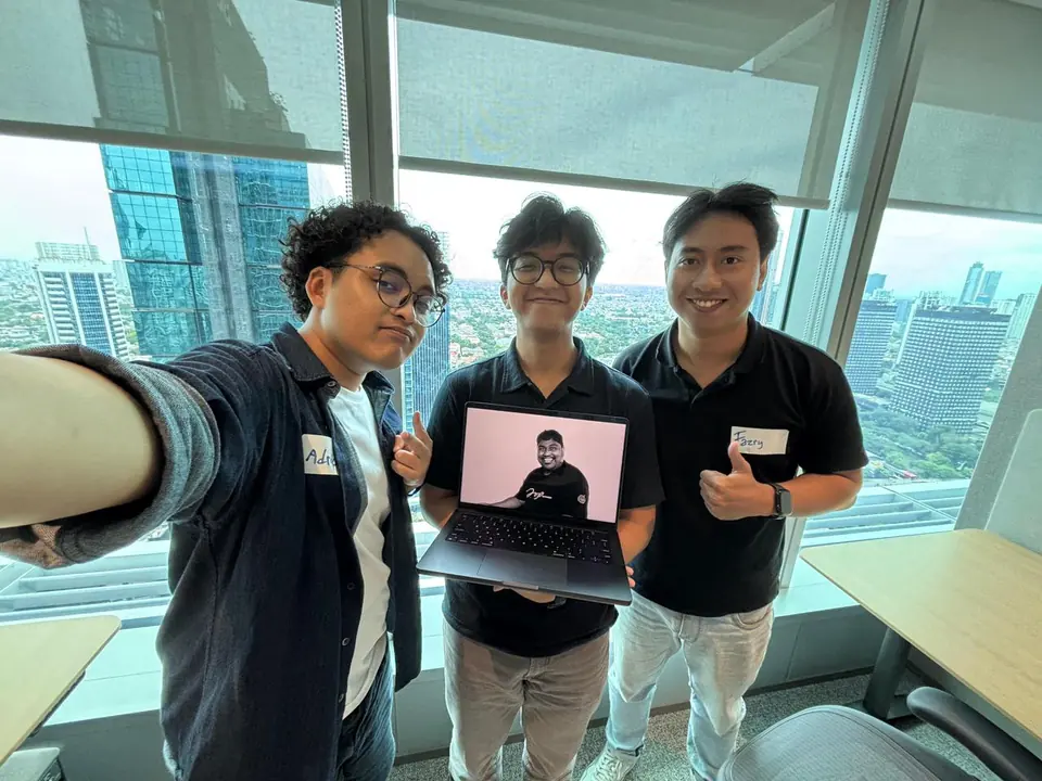 Selfie of Adrian and two teammates by an office window, one holding a laptop showing the absent teammate on a video call