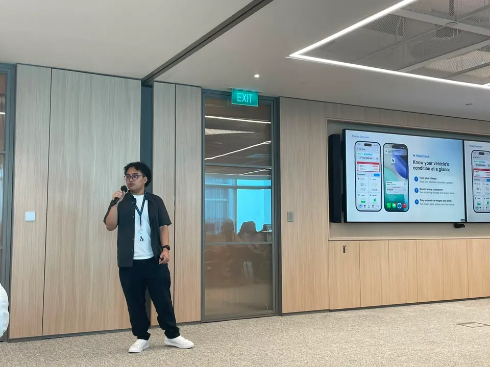Adrian speaking into a microphone in a bright office, app screens on a display behind